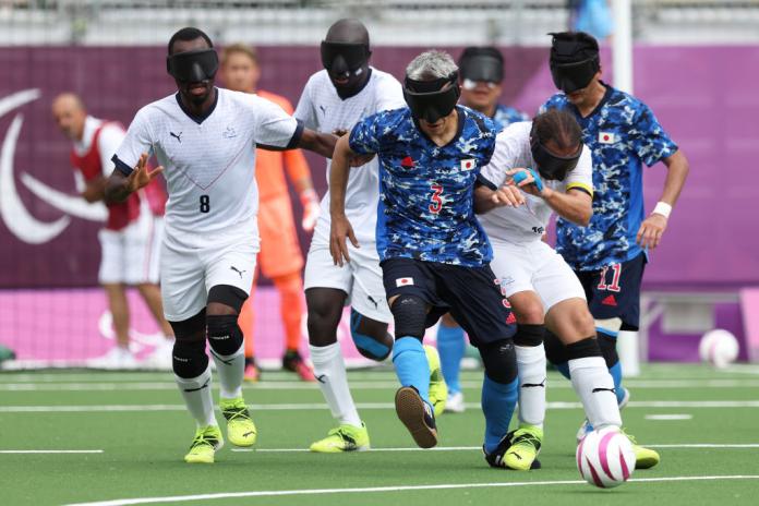 Blind football players in blue and white jerseys compete over the ball at Tokyo 2020.